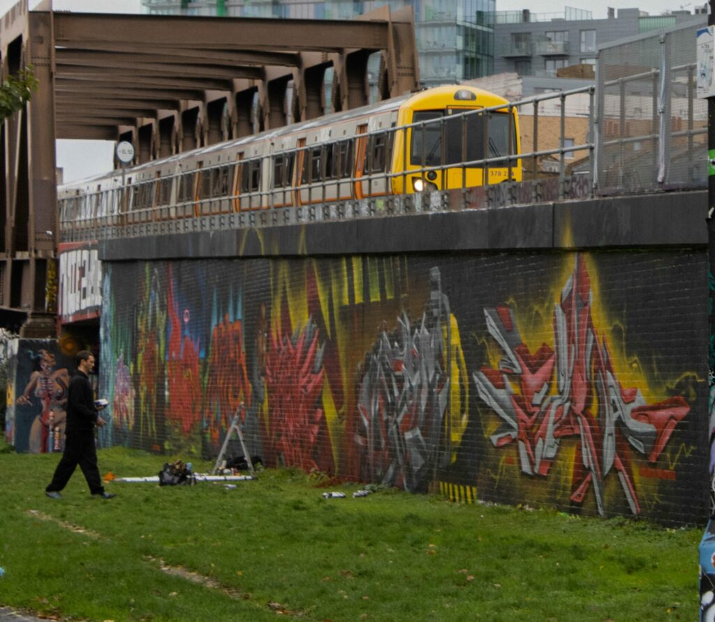 Photo format paysage d'un métro aérien à Londres passant en arrière plan. En avant plan sur la gauche de l'image une personne en train de réaliser un tag.